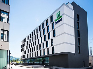 White façade with evenly spaced windows, glass fronts on the first floor. Two shrubs in rectangular planters in front of the entrance