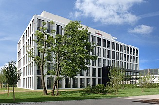 Græsplæne med træer og buske i forgrunden, bagved et hvidt kontorkompleks i fem etager med store vinduer