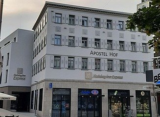 Lys hjørnebygning med bogstaverne Apostel Hof og HolidayInn Express, butiksvinduer nedenunder, smalle vinduer med skodder ovenover