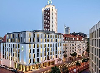 Weißes Eckgebäude an mehrspurigen Straßen, unten Glasfronten zum Foyer, darüber vier Geschosse mit schmalen Fenstern, zwei weitere Geschosse etwas zurückgesetzt darüber. Im Hintergrund Hochhäuser.