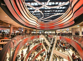 Blick in ein Einkaufszentrum mit zwei Rolltreppen in der Mitte, mehreren Etagen mit Ladenstraßen, die Geländer haben orange-rote geschwungene  Verkleidungeng, die sich an der Decke des Zentrums wiederfinden