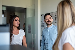 A blonde woman from behind, a man and a woman from the front talking to each other in front of a metaplan wall