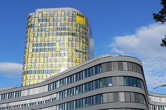 Buet facade i fem etager med vinduer, bagved et tårn med afrundede hjørner og gule farvereflektioner, ADAC-logo øverst, Saturn-skrift over indgangen