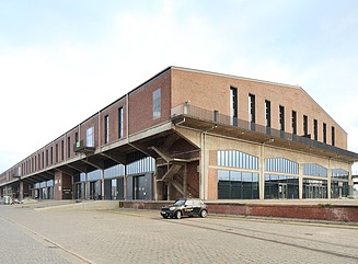 Leicht angerostete Metallgerüste führen zu einem sanierten Lokschuppen aus rotem Backstein, unten mit großen Fensterfronten, oben im Giebel siehr schmale hohe Fenster